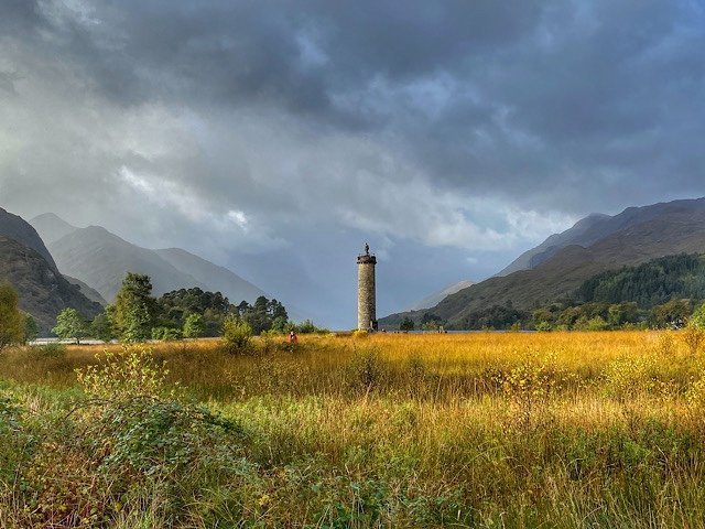 Sunlight on golden grass around the monument of Bonnie Prince Charlie. The loch behind it is surrounded by mountains.