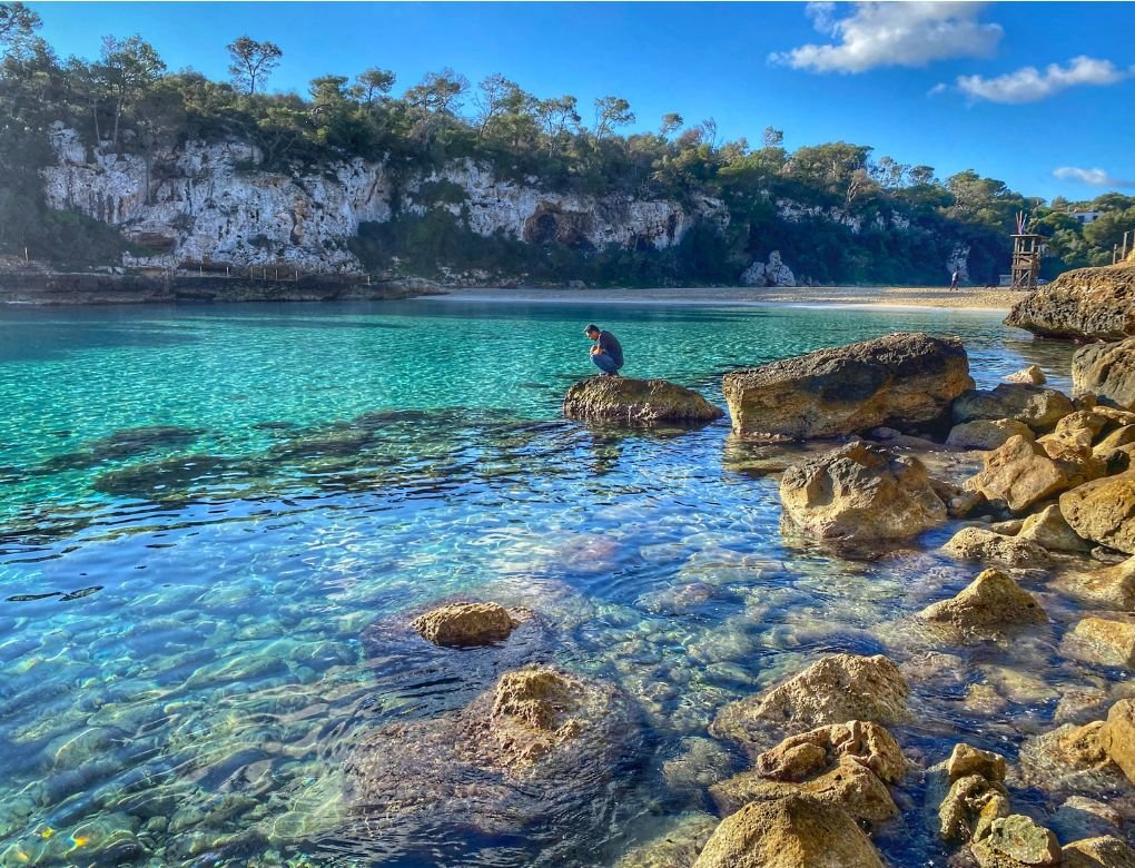 Trin on a rock gazing at the blue waters of a bay in Mallorca