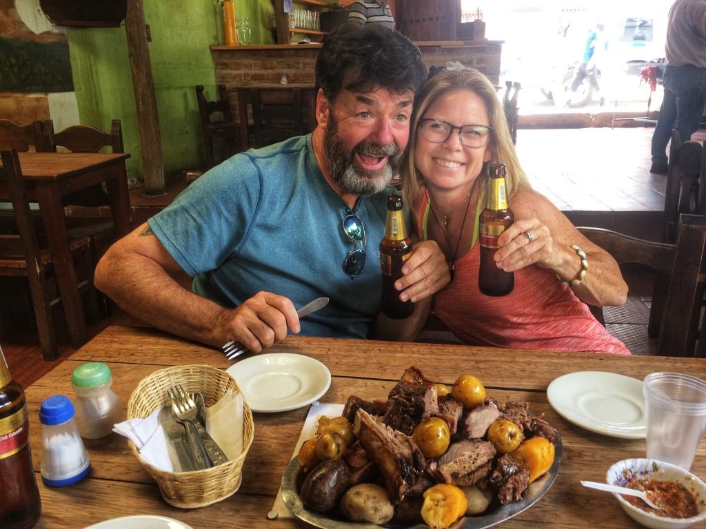 Colin and Sandy posing with their beer at a restaurant. A plate of meat and potatoes sits in the center of the table.