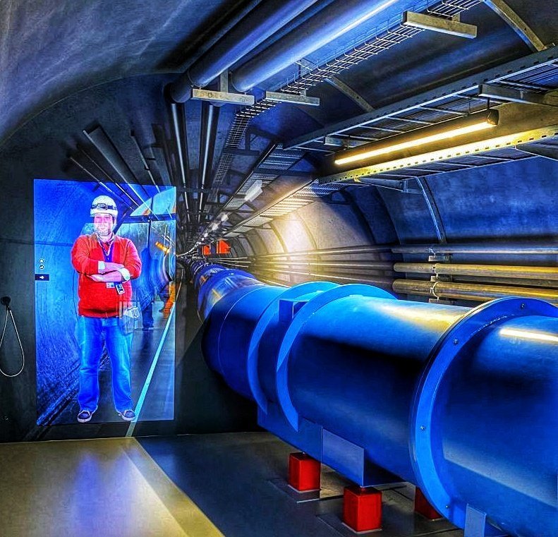 A large blue conduit runs through this mock up of the 27 kilometer tunnel of the LHC. A physicist is projected on the wall and tells about this area of the collider.