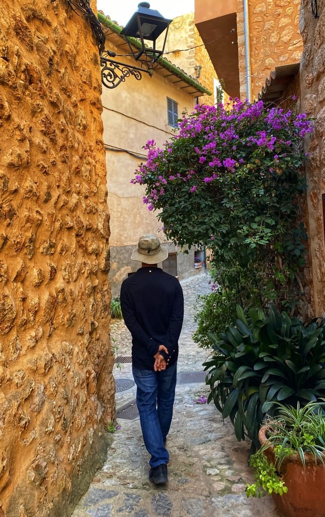 Trin walking down a residential street in Spain. The old walls are made of stone and plants line the narrow walkway on the right.