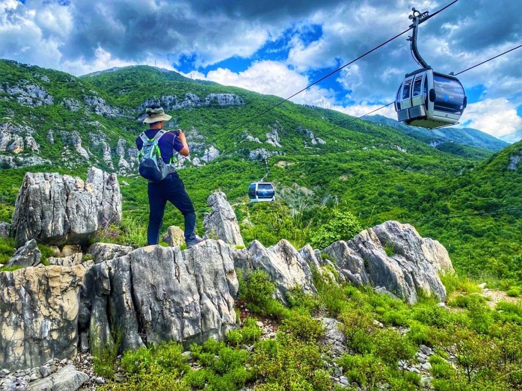 Trin standing on a rocky outcrop that drops off in a cliff on the other side. Cable Cars slide by nearby.