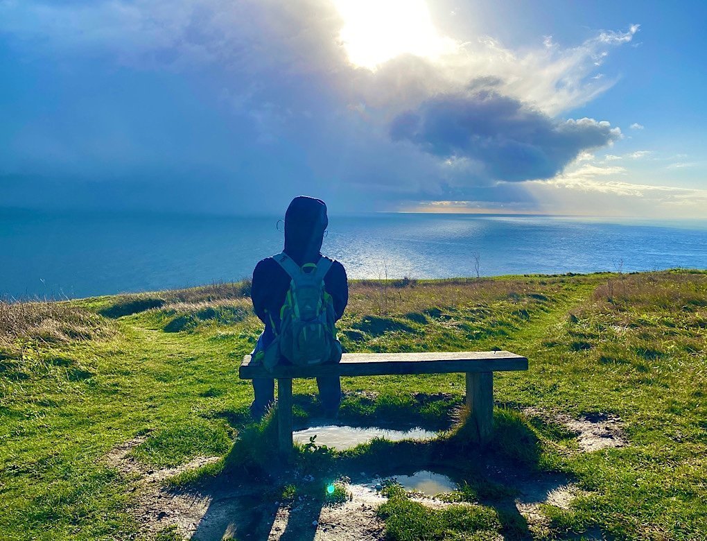 Trin sits on a bench on the Cliffs of Dover looking across the English Channel towards France. The stormy sky is clearing with a bit of sun just appearing.