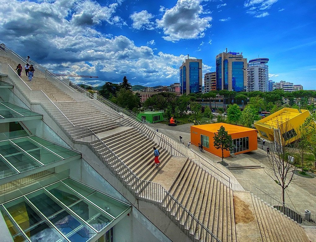 Stairs on the side of the transformed Pyramid of Tiran. Yellow office spaces sit off-kilter beside the pyramid and high rises sit in the background.