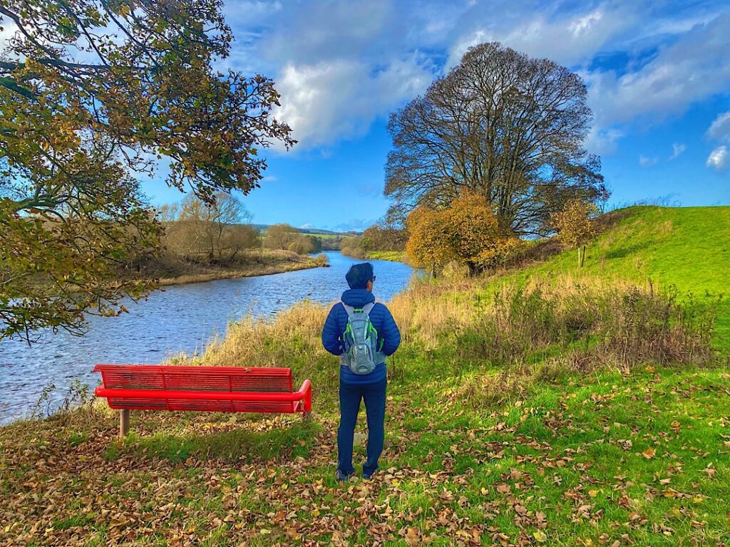 Trin standing by a red bench along the river in Penrith UK