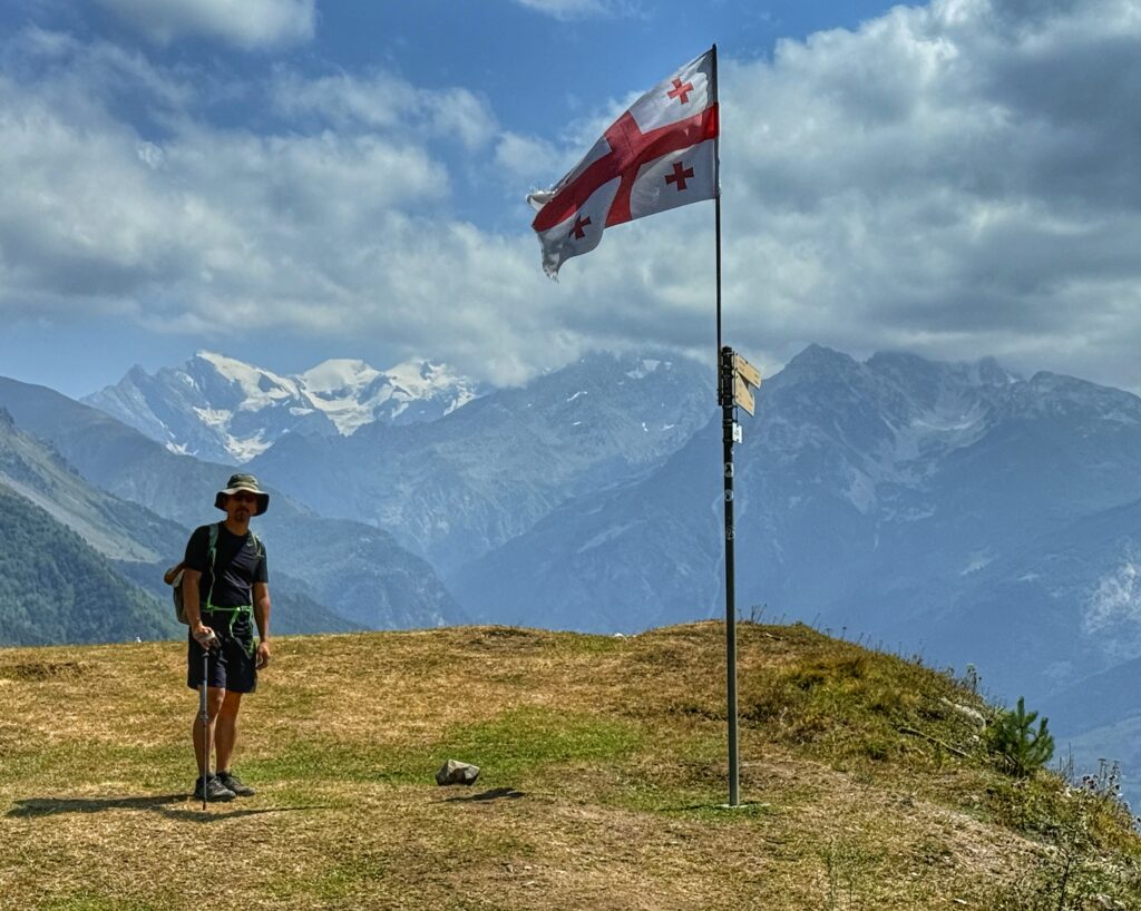 Trin on the mountain pass for day one next to the Georgian flag. Glacier capped mountains are in the background.